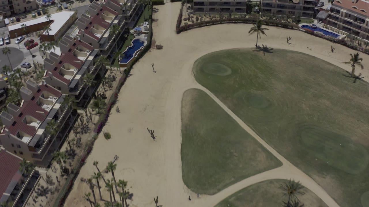 Overhead Los Cabos frame with the open green area centered, pale paths crossing the grounds, buildings tight to the top and right edges, and no visible people.