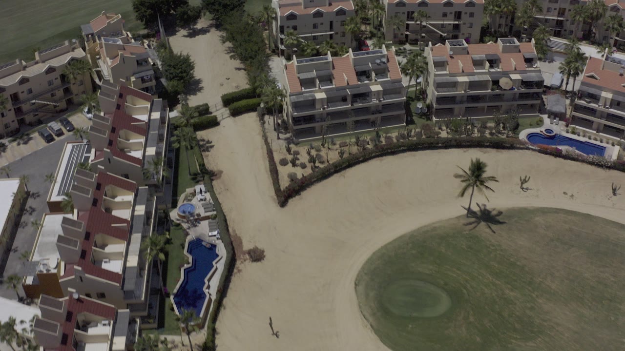 High aerial view in Los Cabos, Mexico showing vacation rental buildings around sand-colored paths, small blue pools, palms, and golf-course grass, with no visible people.