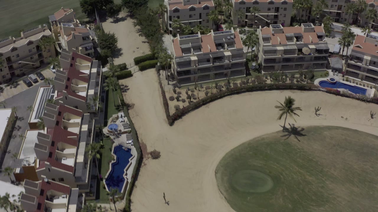 Aerial frame in Los Cabos, Mexico looking down on tan rental buildings, red roof sections, a curved sand area, palms, pools, and nearby golf turf, with no visible people.
