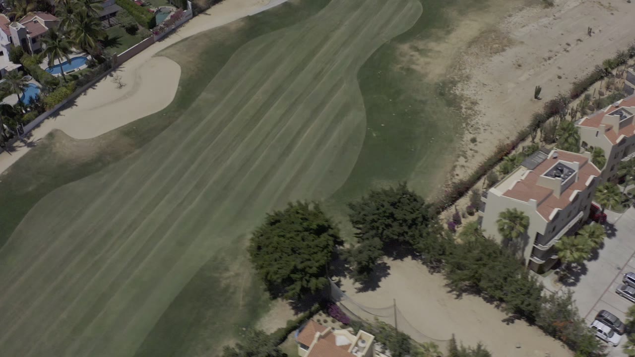 Aerial frame in Los Cabos, Mexico with the fairway running diagonally past a sand bunker, palm-lined houses, blue swimming pools, and no visible people.