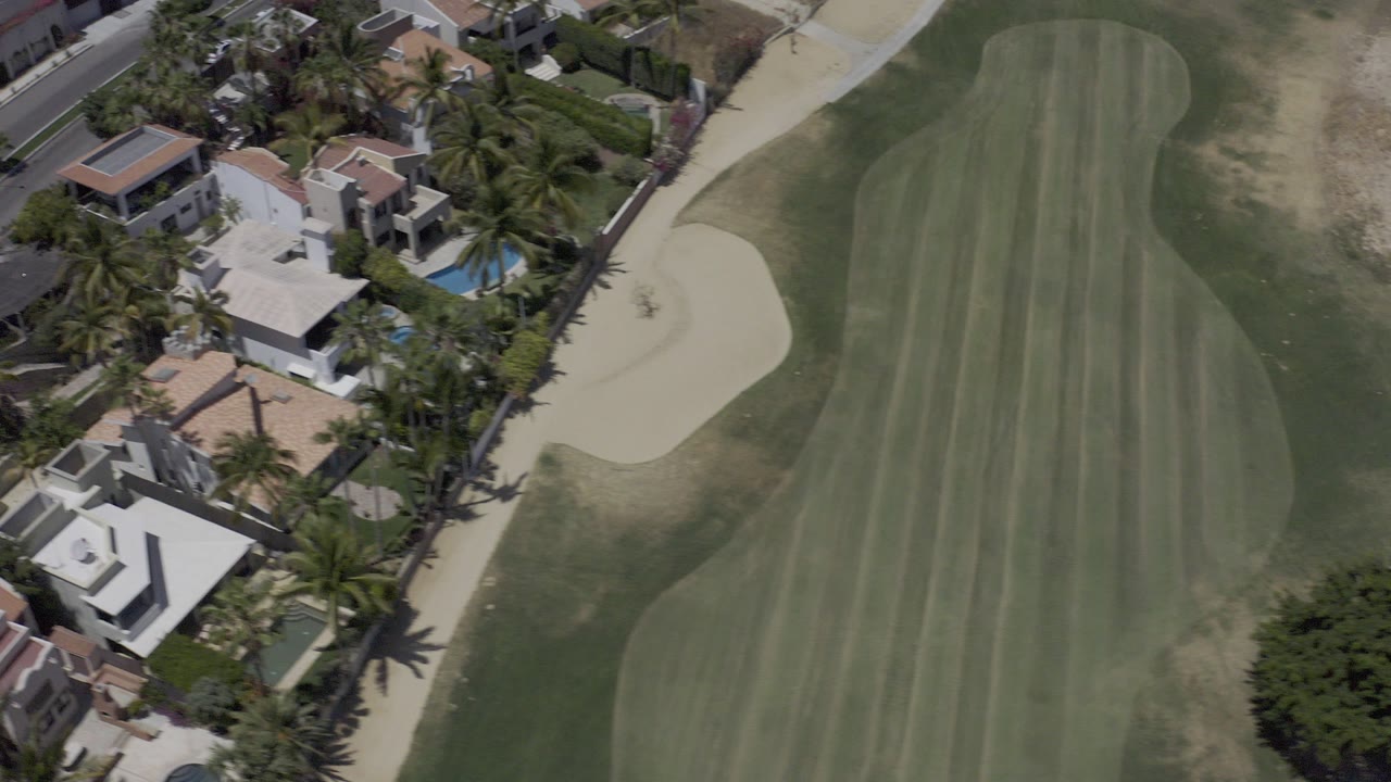 High aerial frame over Los Cabos showing a striped golf fairway beside pale sand bunkers, white homes, palm landscaping, and backyard pools, with no visible people.