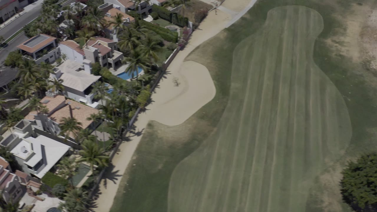 Los Cabos aerial frame with more residential roofs and blue pools visible beside the fairway and a large sand bunker, with no visible people.