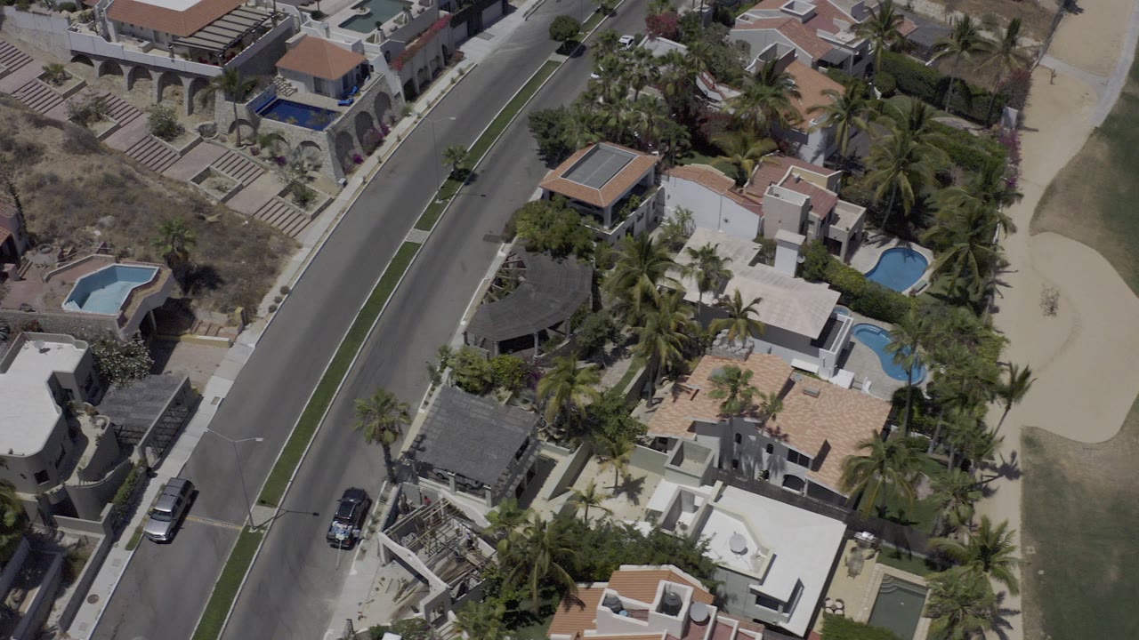 Overhead aerial frame in Los Cabos, Mexico showing luxury homes, blue private pools, palm trees, dry hillside ground, a diagonal road, and no visible people.