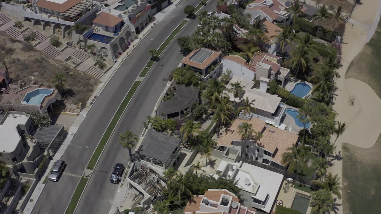 Aerial frame in Los Cabos, Mexico with the diagonal street centered between upscale houses, walled yards, blue pools, palms, cars, and no visible people.