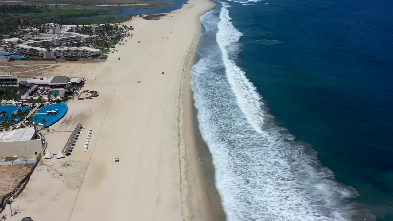 Aerial beach view with resort buildings, pale sand, and breaking surf.