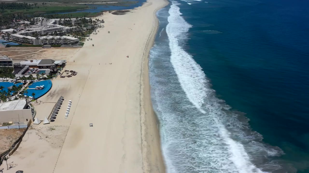 High aerial view in Los Cabos with a long sandy beach, foamy surf moving along the waterline, beachfront buildings on the left, and no visible recognizable people.
