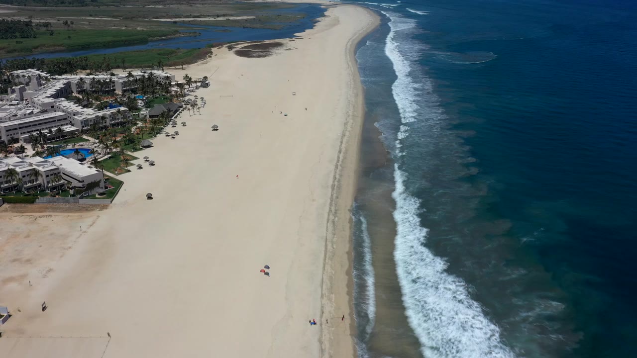 High aerial view over a Los Cabos beach, with resort buildings and pools at left, pale sand through the center, blue ocean at right, and no clearly visible people.