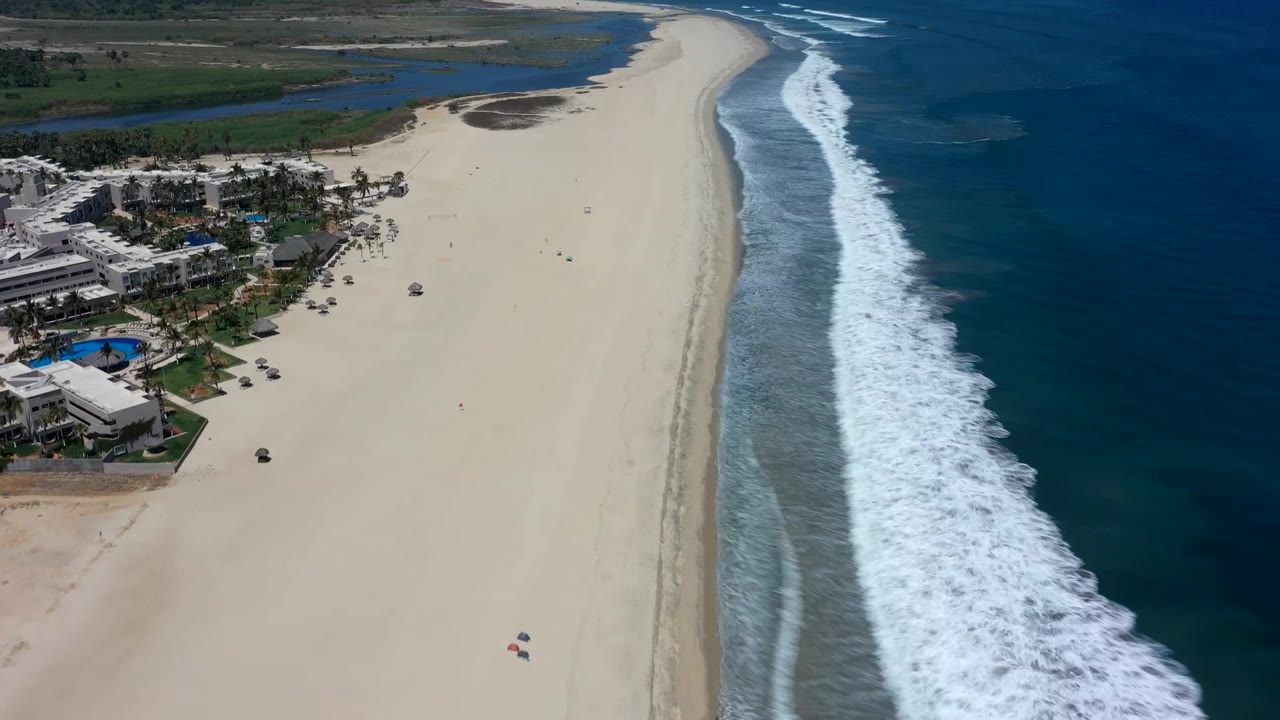 Wide aerial beach scene with ocean surf and buildings near the sand.