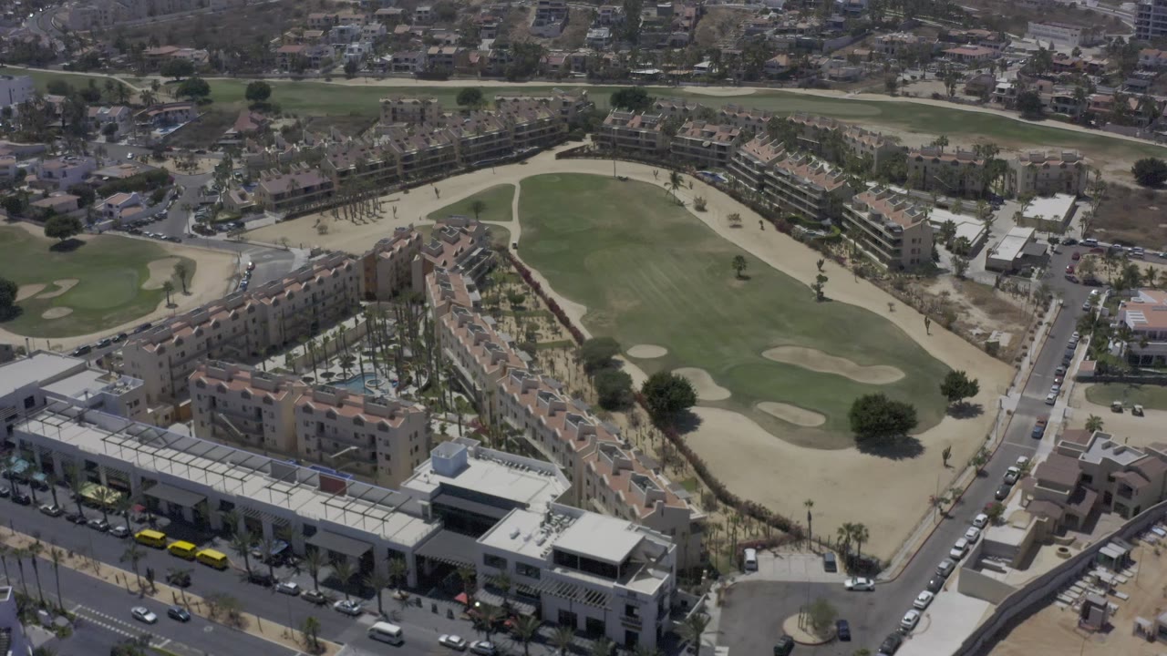 A Los Cabos drone-like view centers the green course area, with apartment-style buildings and a palm-lined courtyard in the foreground and no visible people.