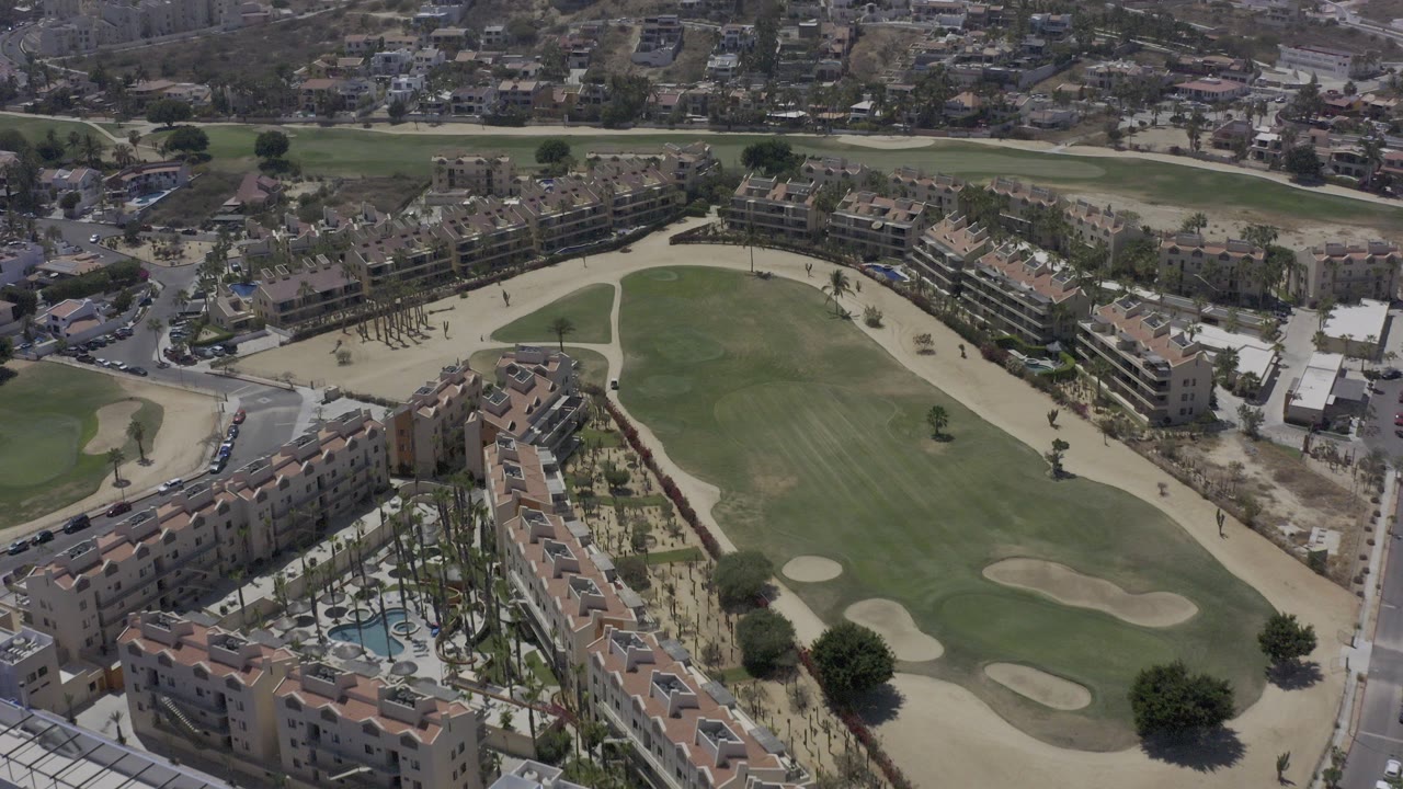 Aerial frame from Los Cabos, Mexico, showing tan-roofed vacation rental buildings, palm-lined courtyards, a green golf fairway, pale bunkers, surrounding roads, and no visible people.