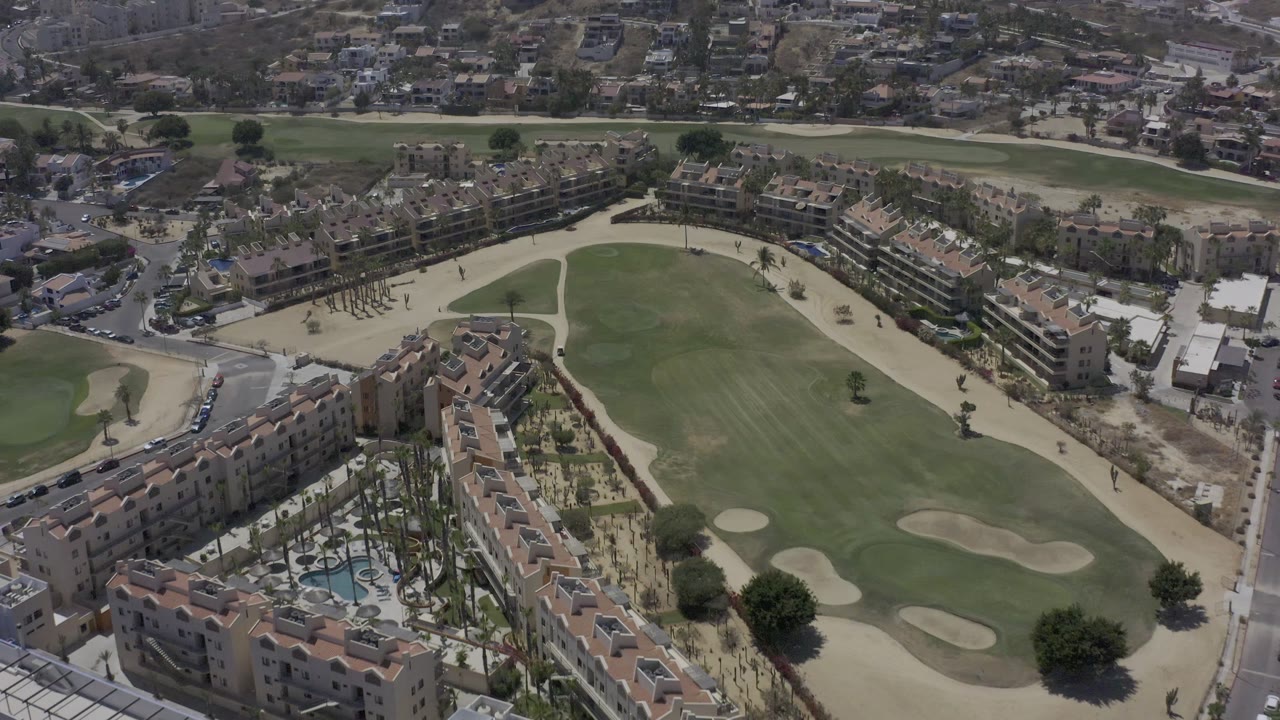Aerial view in Los Cabos, Mexico, with the rental buildings filling the lower frame, pool areas and palms visible inside the courtyards, the golf course across the center, and no visible people.