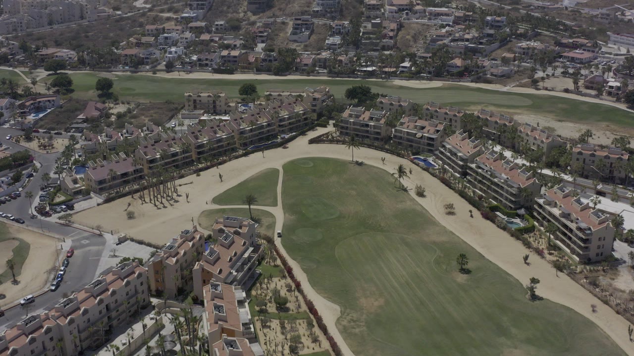 Aerial view in Los Cabos, Mexico, showing a green field below terracotta-roofed buildings, pale paths, roads, parked cars, and hillside housing with no visible people.