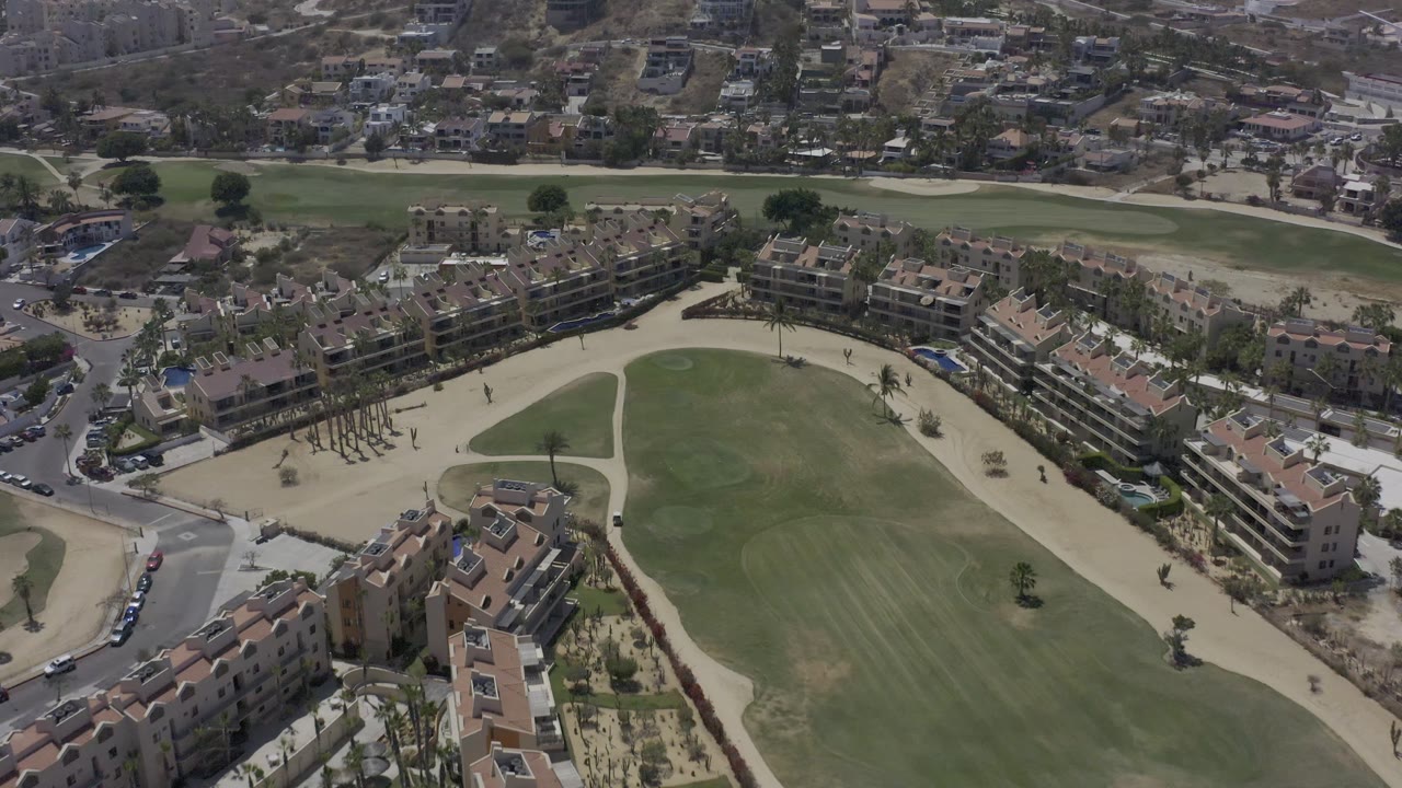 High aerial frame in Los Cabos, Mexico, with the field centered between curved paths, apartment-style buildings, nearby roads, parked cars, and distant hillside homes with no visible people.