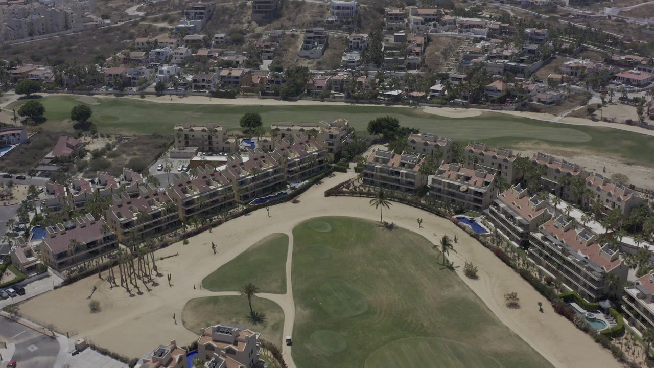 High aerial view in Los Cabos showing tan-roofed residential buildings, green fairways, pale sand areas, and hillside housing in the background with no visible people.