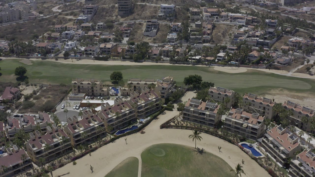 High aerial view over Los Cabos, Mexico, with tan-roofed condos in the foreground, a green golf fairway beyond them, hillside housing in the distance, and no visible people.