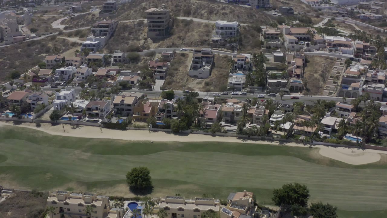 Aerial view in Los Cabos, Mexico, with sand bunkers, green turf, roadside homes, and sunlit dry slopes in the background, with no visible people.