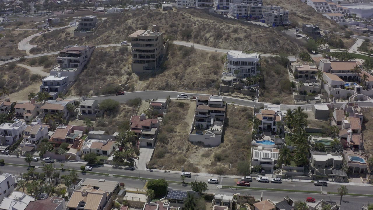 Aerial frame over Los Cabos hillside homes and dry scrub lots, with a curved street and small cars visible below, no visible people.