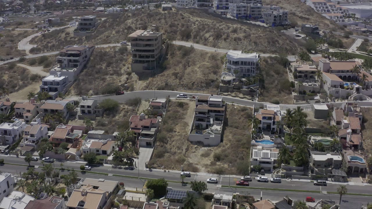 Elevated Los Cabos view with separated houses, pale dry terrain, parked cars along the street, and larger buildings on the hillside, no visible people.