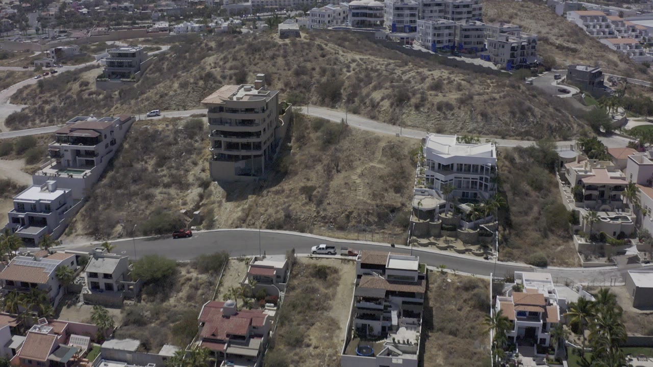 Aerial frame from Los Cabos, Mexico showing dry hillside lots, several modern homes, a curved road, and no visible people at the start of the clip.
