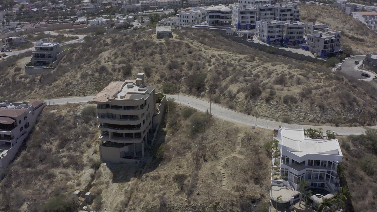 Aerial view over Los Cabos hillside homes, dry lots, sparse shrubs, and winding roads, with no visible people.