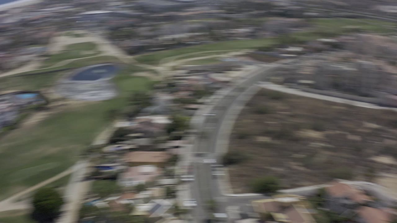 Second blurred aerial frame over Los Cabos residential streets, green grounds, sandy paths, and the coastline beyond, with no visible people discernible.