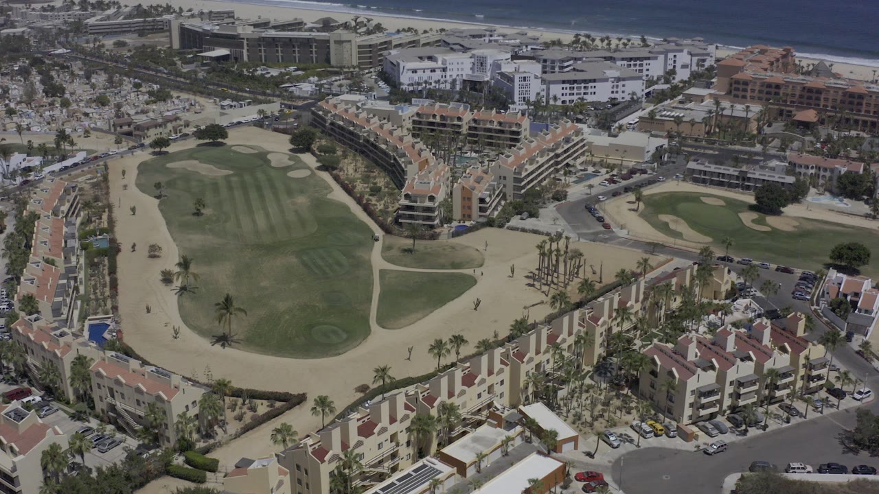 Opening aerial frame over Los Cabos, Mexico, showing red-roof condo buildings, roads, green open ground, sandy paths, and a beach and ocean strip beyond, with no visible people.
