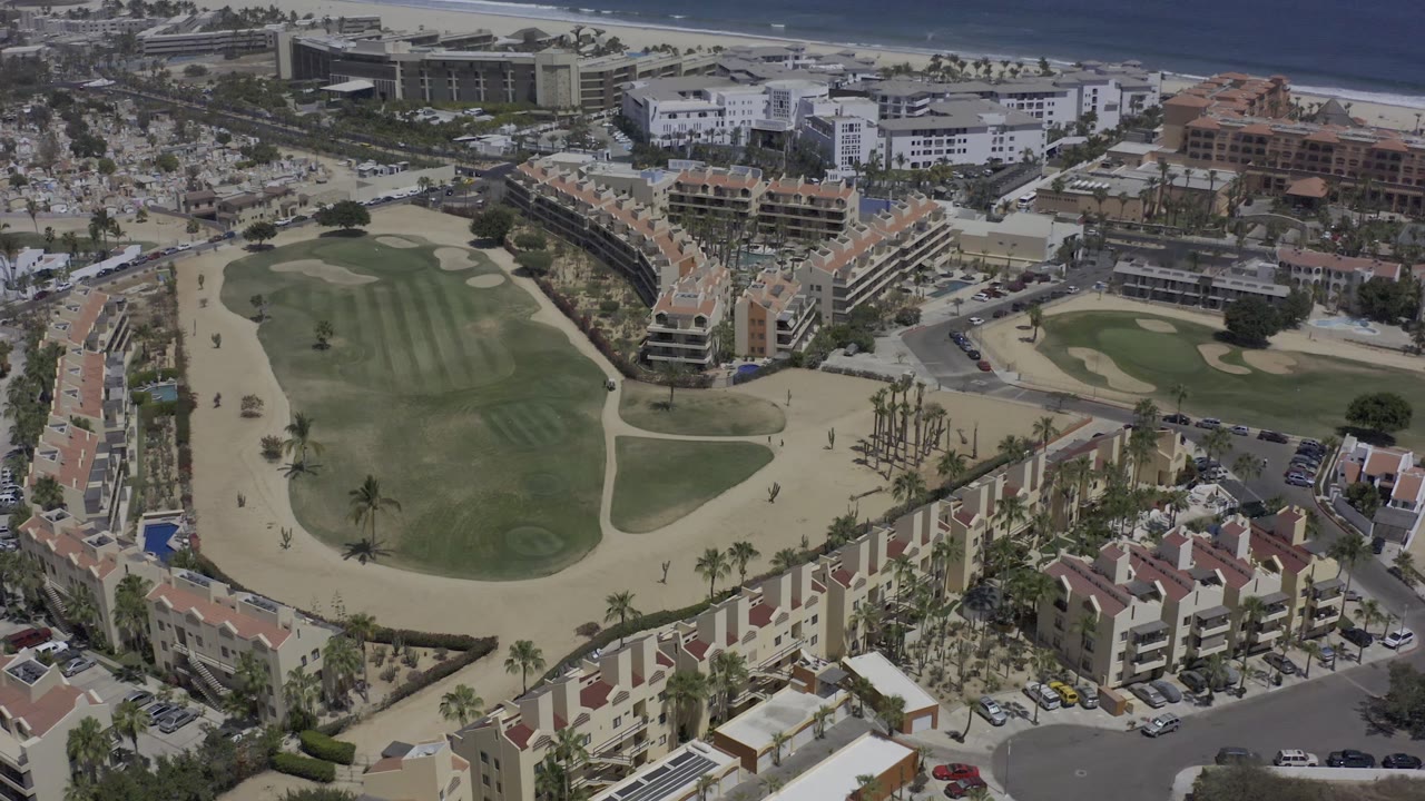 Second aerial frame over Los Cabos, Mexico, with the central green space and sand paths framed by condo blocks, parking areas, palm trees, and the shoreline at the top, with no visible people.