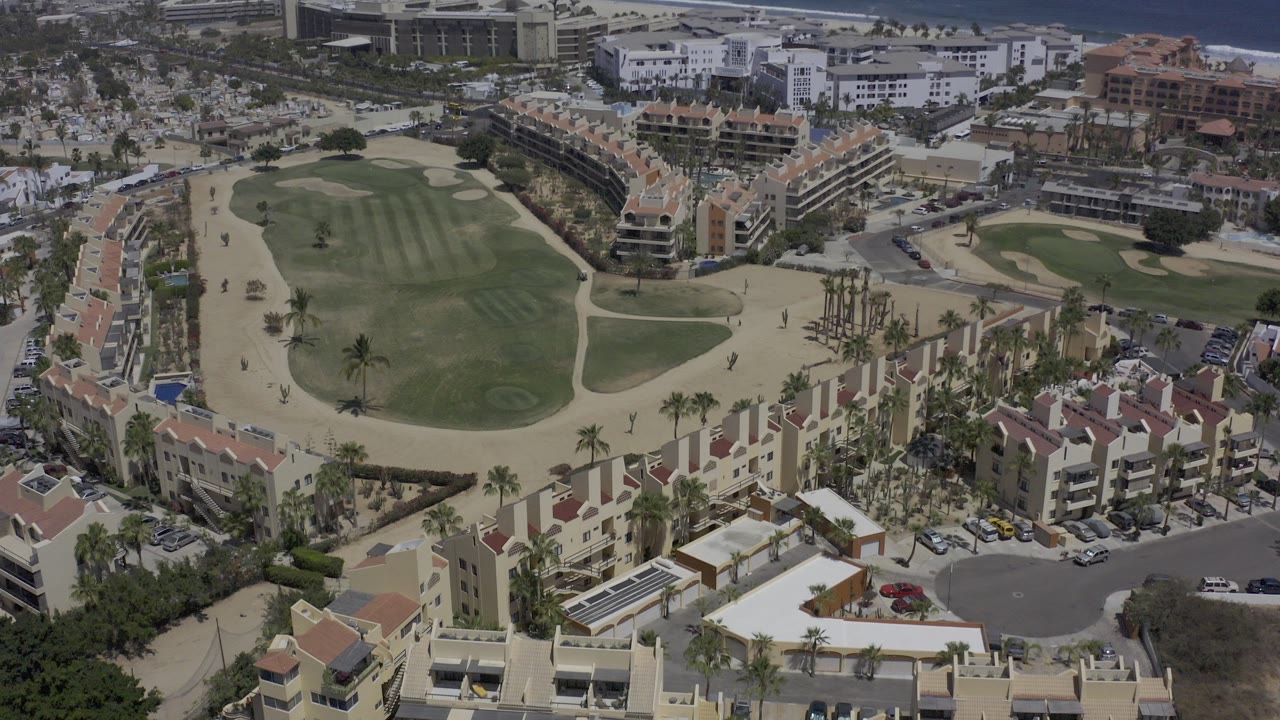 Aerial view over condos, a green fairway, sandy paths, and distant coastal buildings.