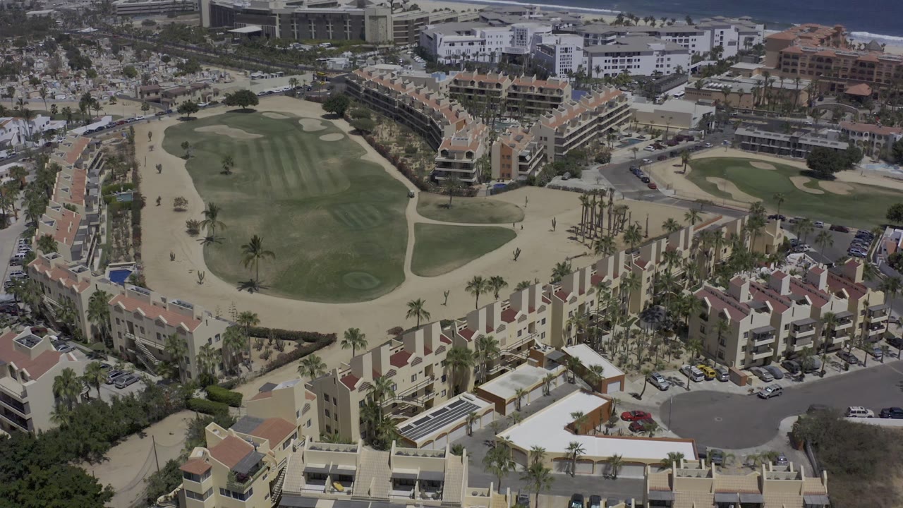 A Los Cabos aerial still centers the green fairway and pale walking or cart paths, with condo blocks along both sides, streets beyond, and no visible people.