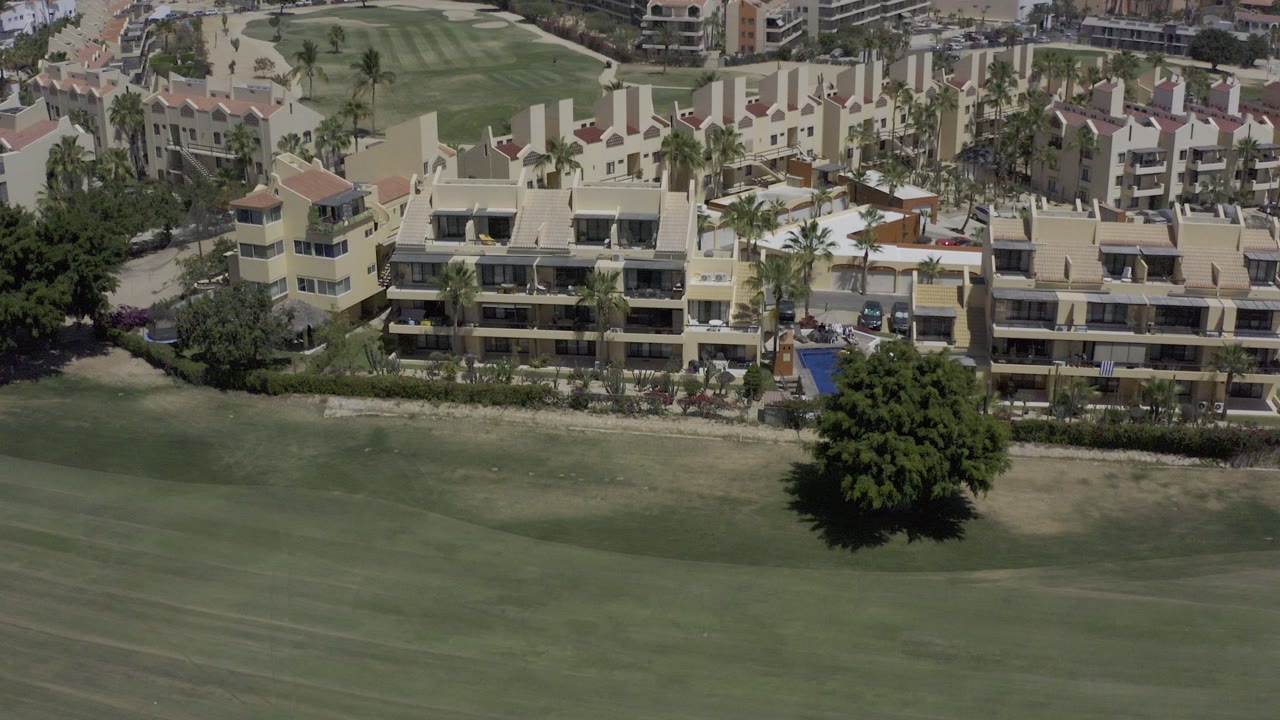 Opening aerial frame over a Los Cabos golf course, with wide green fairway in front, cream-colored residential buildings behind it, palm trees, and no visible people.