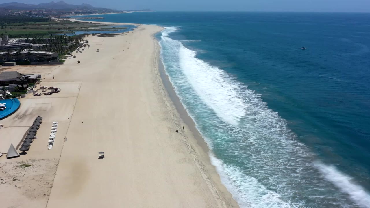High aerial view over a Los Cabos beach, with pale sand on the left, white surf running down the shoreline, blue water to the right, and no clearly visible people.