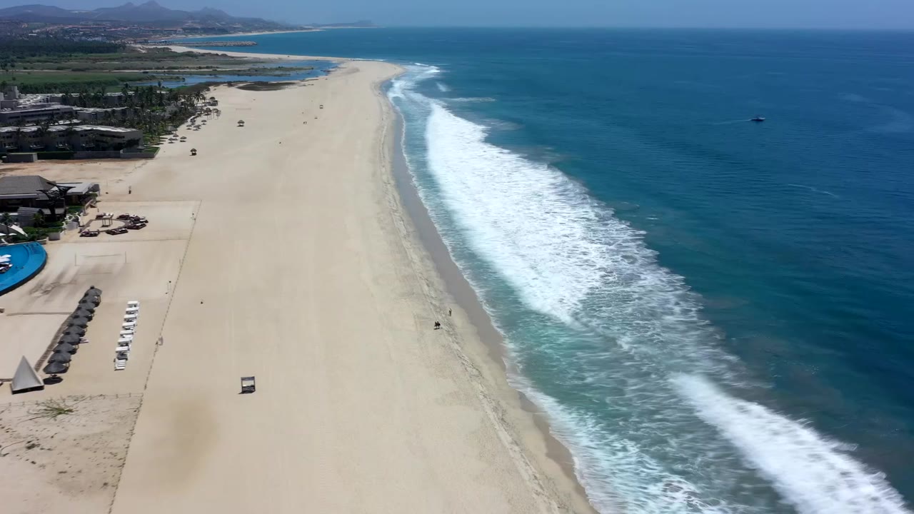 Aerial Los Cabos shoreline view showing the beach stretching toward distant hills, a bright surf line near center, open ocean on the right, and no clearly visible people.