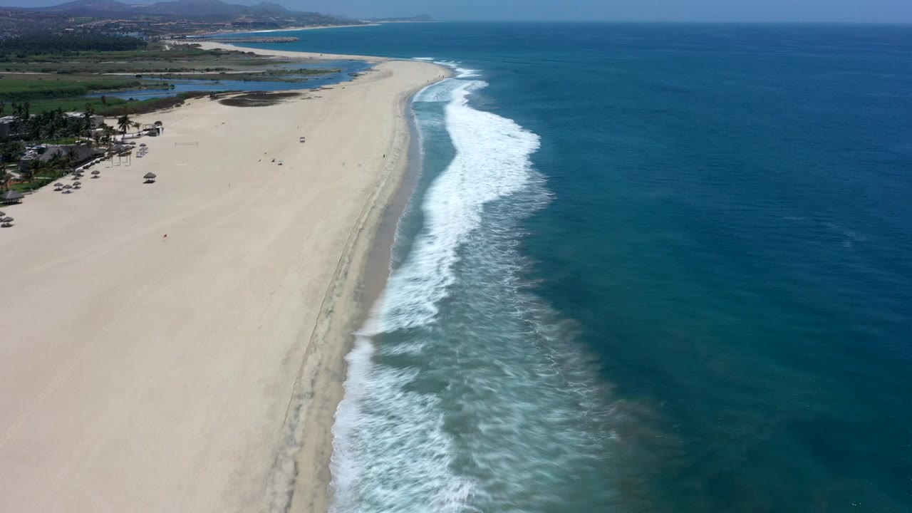 Aerial view of a long sandy beach with waves breaking beside deep blue water.