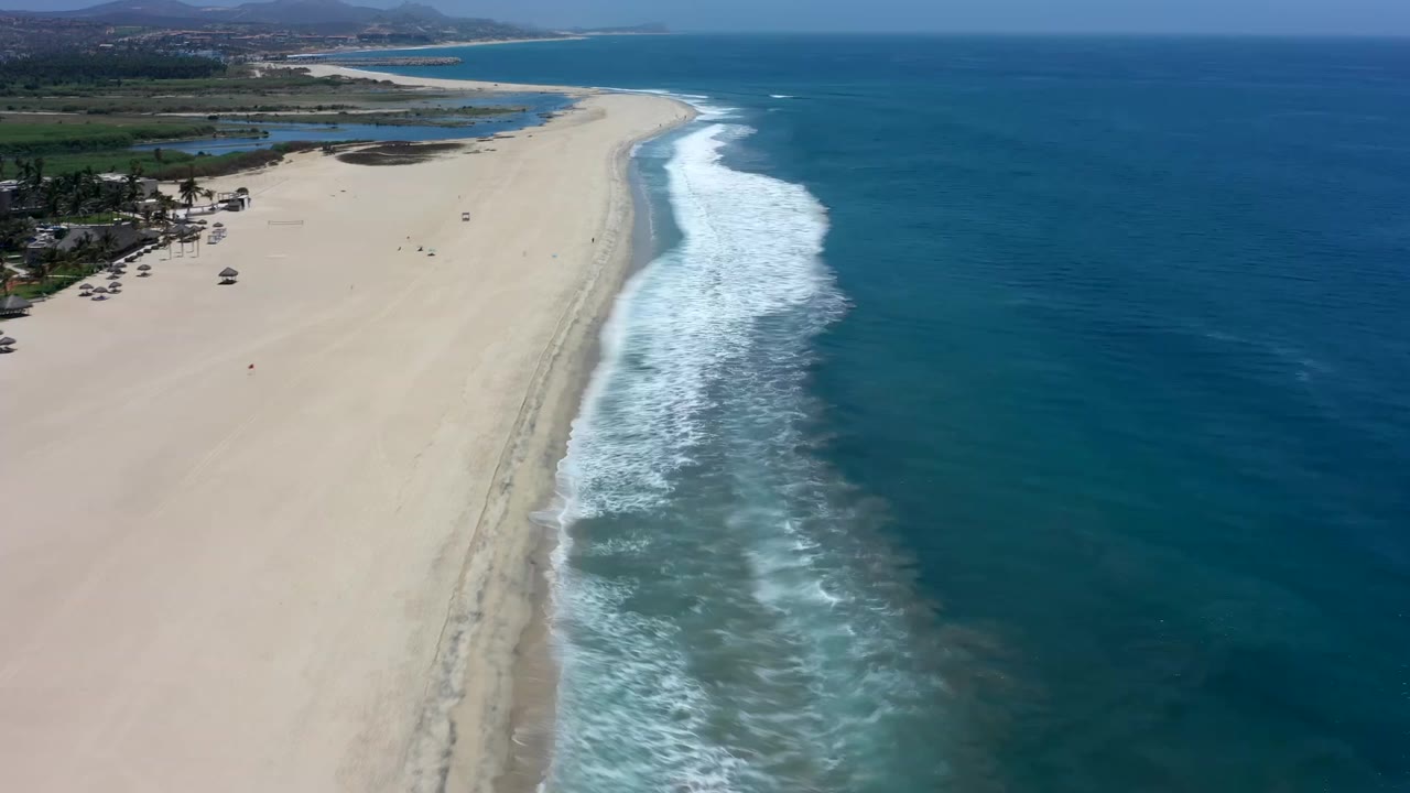 High view of the Los Cabos shoreline with foamy waves pushed farther up the sand, blue water filling the right side, and small people scattered on the beach.