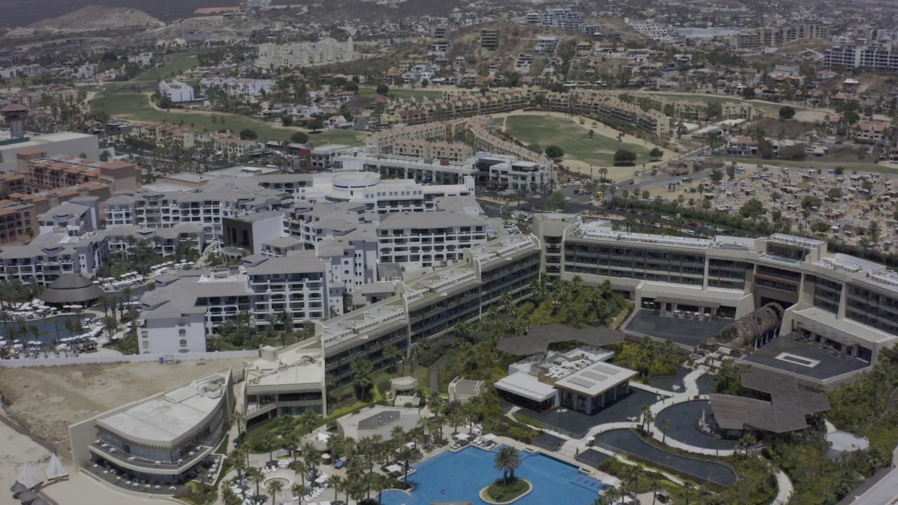 Aerial frame in Los Cabos, Mexico, showing the main white building block, nearby pool decks, a dark-roofed structure, roadways, golf-course turf, distant hillside buildings, and no visible people.