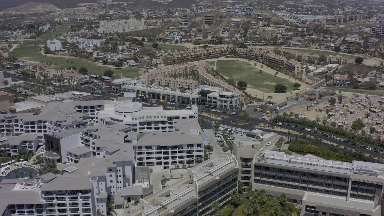 Opening aerial frame over Los Cabos, Mexico, with white multi-story buildings in the foreground, roads and golf greens beyond, hillside development in the distance, and no visible people.