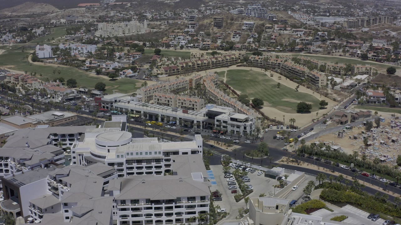High aerial frame over Los Cabos, Mexico with white buildings in the foreground, a road grid, a golf course, hillside neighborhoods, and no visible people.