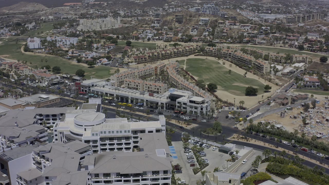 Los Cabos aerial frame looking over foreground office-style buildings, a divided road with vehicles, a green fairway, tan construction ground, and no visible people.
