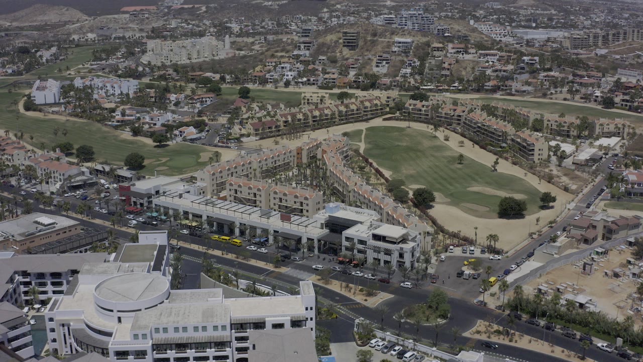 Wide aerial frame over Los Cabos, Mexico, showing white commercial buildings in the foreground, a road junction, rows of homes, and a green golf course beyond, with no clearly visible people.