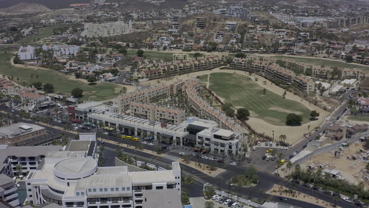 Aerial view in Los Cabos, Mexico, with flat-roofed office buildings and parking lots in front, a long row of homes at center, and golf-course fairways stretching behind them, with no clearly visible people.