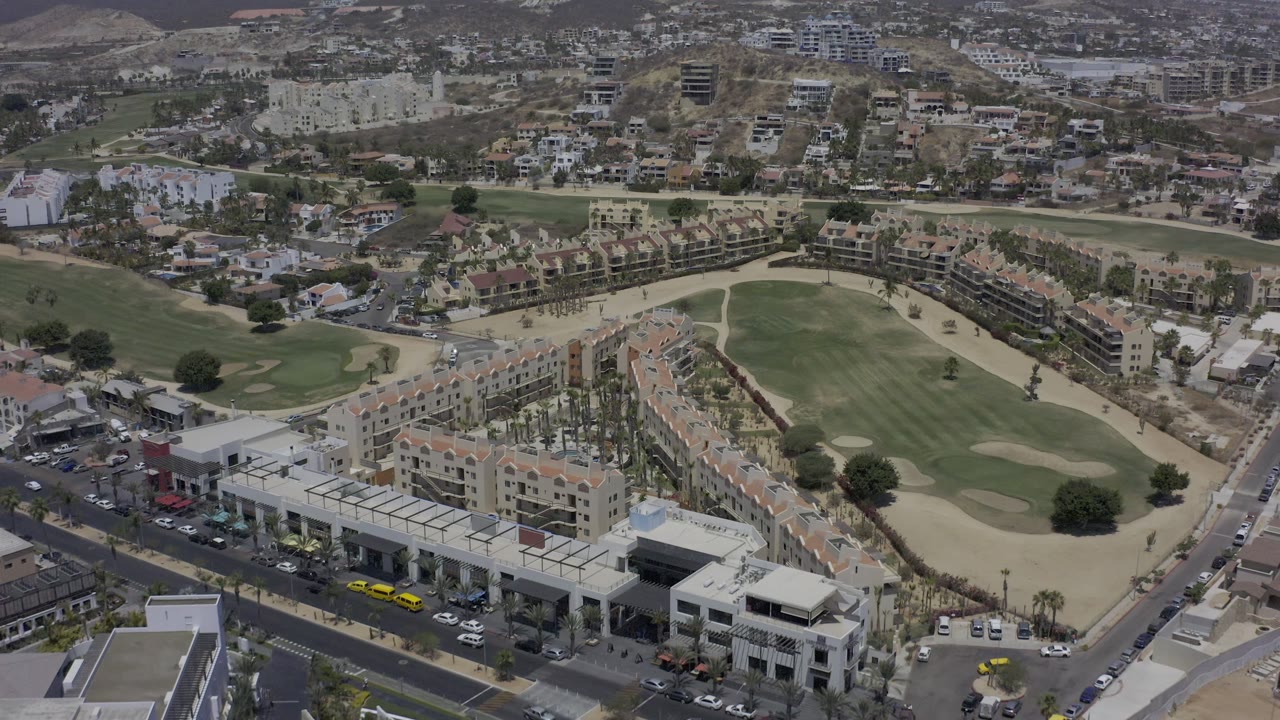 Aerial frame over Los Cabos, Mexico, with the golf course centered, apartment blocks in the lower foreground, street traffic lanes, and no visible people.