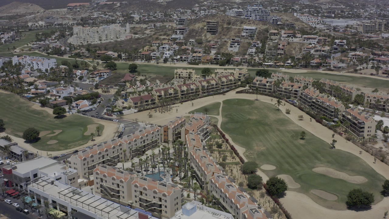 High aerial view over Los Cabos with orange-roofed apartment blocks in the foreground, golf course fairways and bunkers through the center, hillside development beyond, and no visible people.