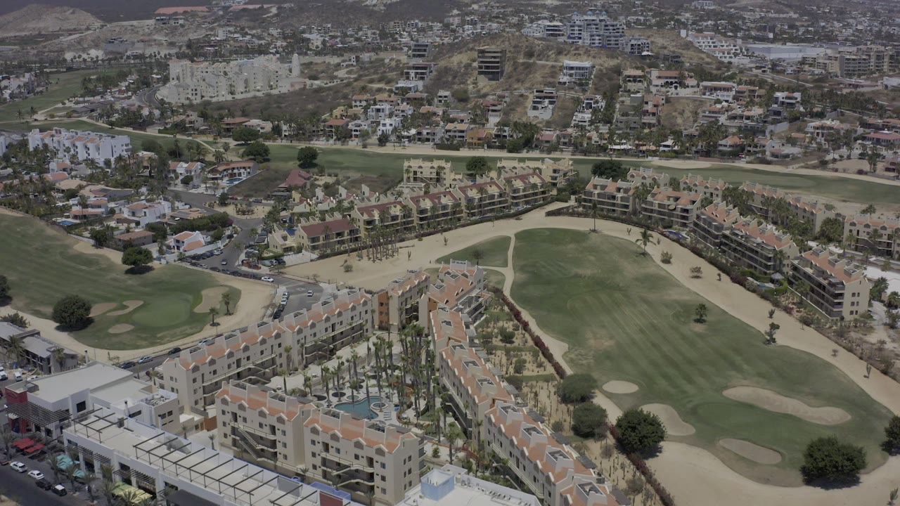 Los Cabos aerial frame with tan residential blocks along the lower edge, a green golf fairway and pale bunkers in the middle, roads curving between buildings, and no visible people.