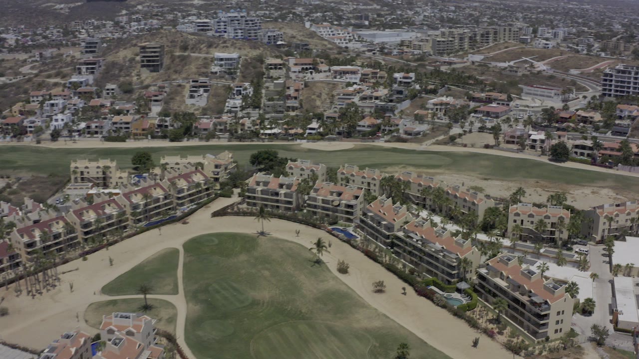 Aerial view of tan-roofed buildings beside green golf course grounds and hillside homes.