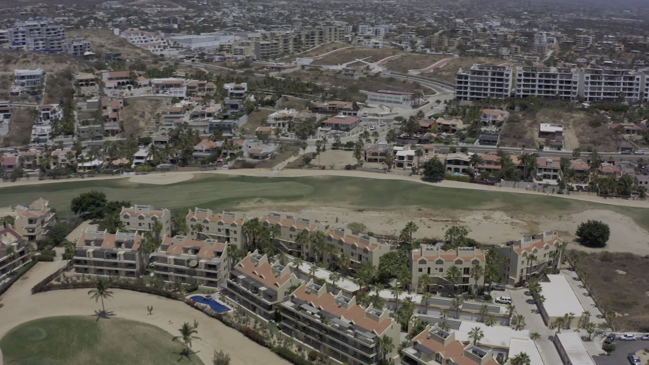 Los Cabos aerial frame with tan-roofed residences in the lower frame, a green fairway through the middle, and larger hillside buildings beyond, with no visible people.