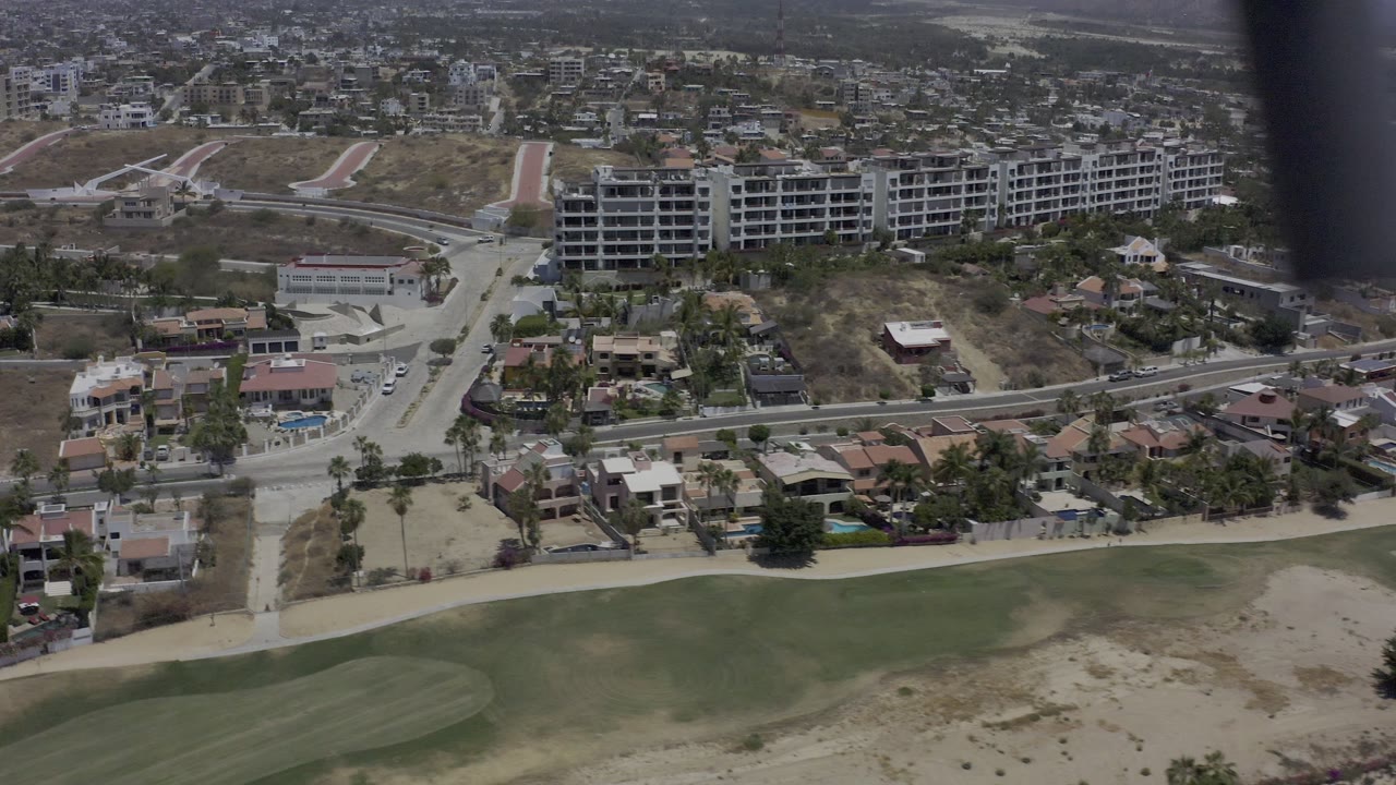 Aerial view in Los Cabos, Mexico showing a road junction, red-roof houses, dry undeveloped lots, larger buildings, and no visible people.