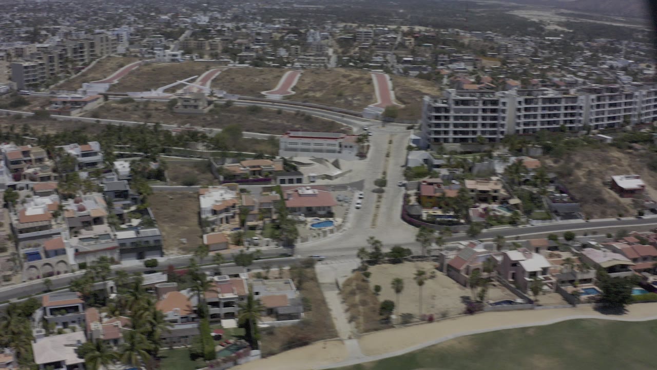 Elevated aerial frame over Los Cabos homes, a curving road, palm trees, dry lots, and larger buildings in the distance, with no visible people.