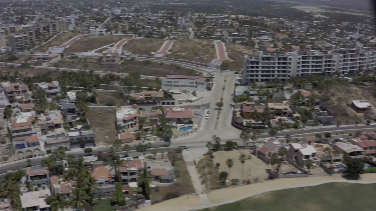 A second elevated view of Los Cabos residential blocks shows villas, dry hillside lots, a main road, and mid-rise buildings under hazy daylight, with no visible people.