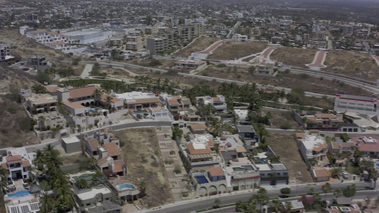 High aerial view over Los Cabos hillside homes, roads, modern buildings, dry grass, and pools in bright daylight, with no visible people.