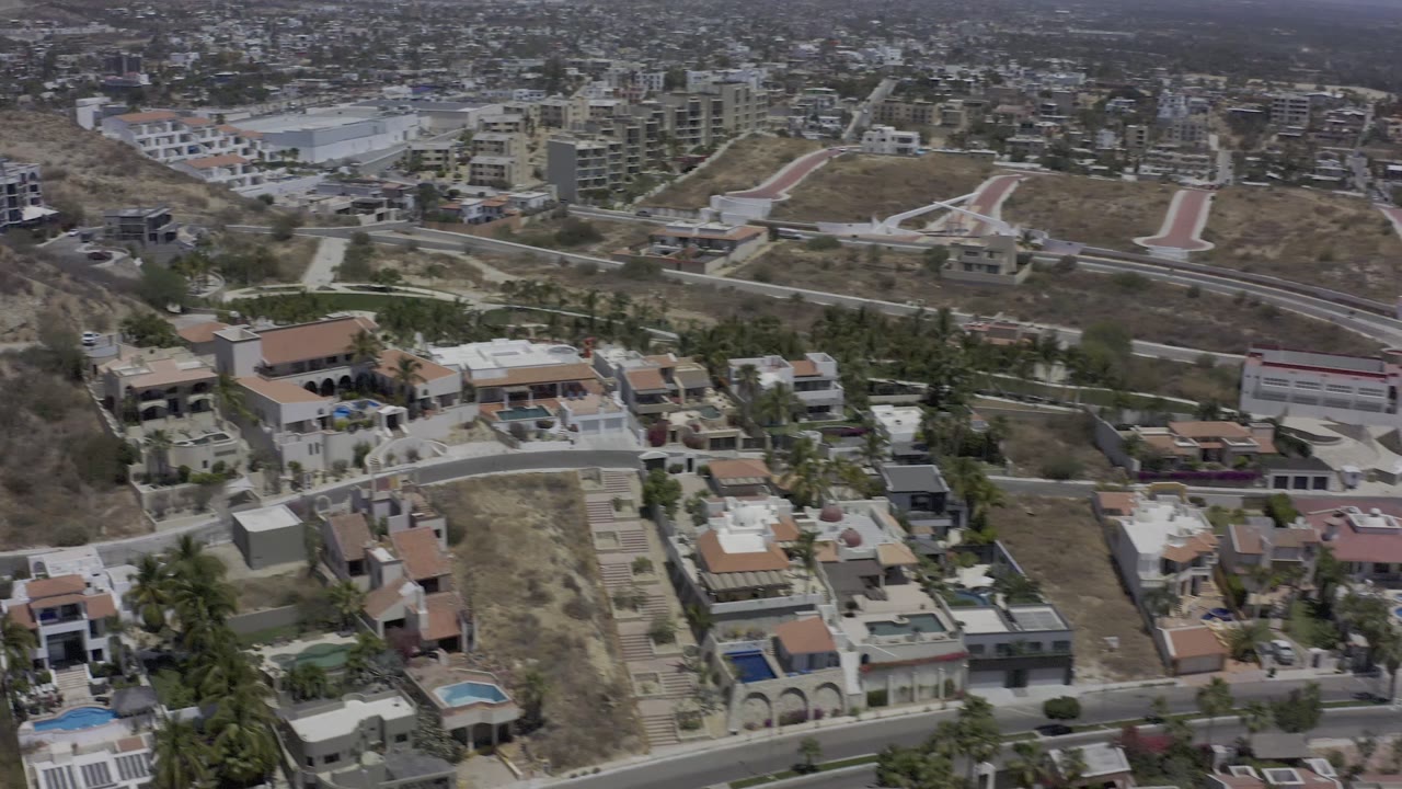 Los Cabos aerial frame looking across large residences, terraced dry hills, curved roads, and distant urban buildings, with no visible people.